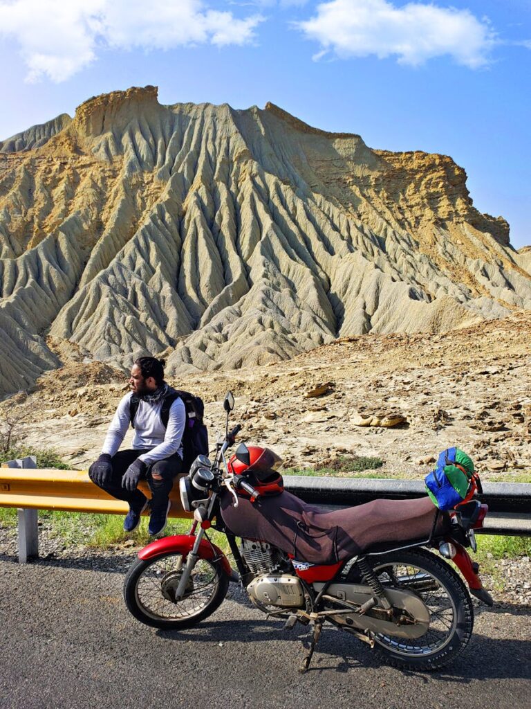 A traveler pauses by a motorcycle with Balochistan's dramatic landscape in the background.